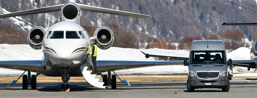 Ground crew member in hi-vis at the door of a business jet on a snowy alpine apron, with a support van alongside